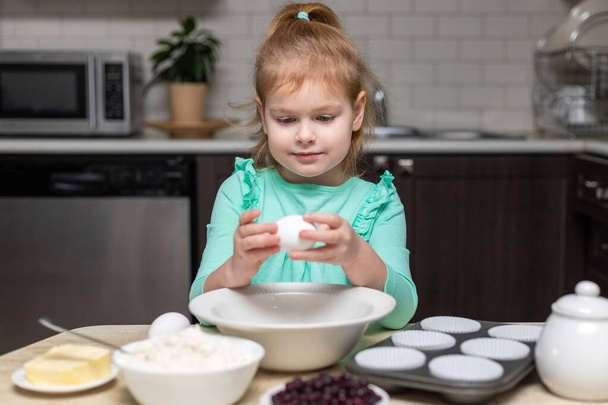 Child cooking muffins in the kitchen. Ingredients for baking on table. Little cute girl ready to break an egg to make pastry - Foto, Imagem