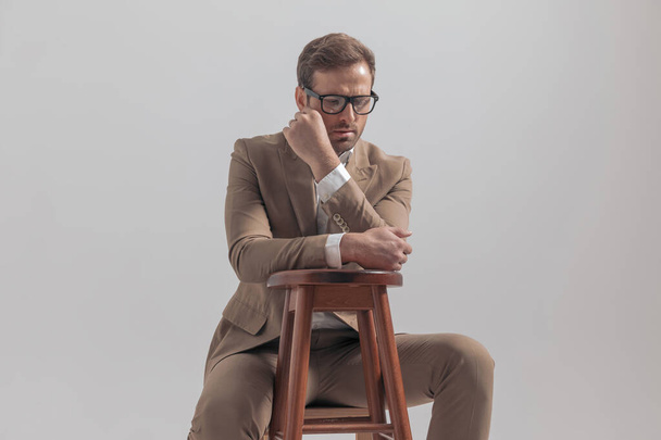 atractivo hombre de negocios sentado en una esquina y siendo dramático, mirando hacia abajo contra el fondo gris - Foto, imagen