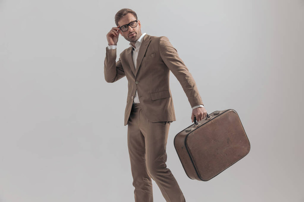 boczny widok seksownego biznesmena układającego okulary i trzymającego walizkę na szarym tle - Zdjęcie, obraz