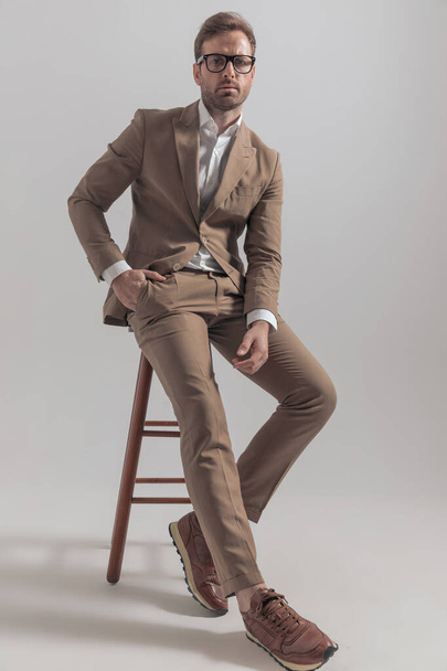 sexy macho uomo d'affari in posa sulla sedia con atmosfera fresca, indossando occhiali da vista e un abito marrone - Foto, immagini