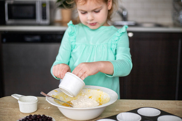 Ένα παιδί μαγείρευε κεκάκια στην κουζίνα. Κορίτσι ανάμειξη igredients για το ψήσιμο σε μπολ. Παιδική ζύμη, προσθήκη αλεύρου. - Φωτογραφία, εικόνα
