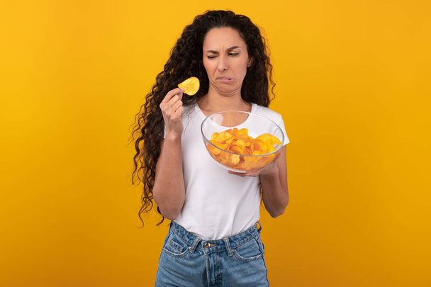 Verwirrte junge Frau isst unappetitliche Kartoffelchips - Foto, Bild