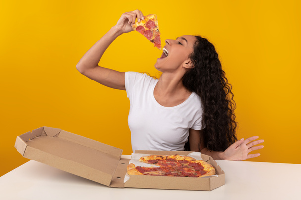 幸せなラテン語の女性がテーブルに座ってピザをかむ - 写真・画像