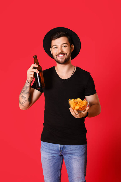 Young man with bottle of beer and chips on red background - Fotó, kép