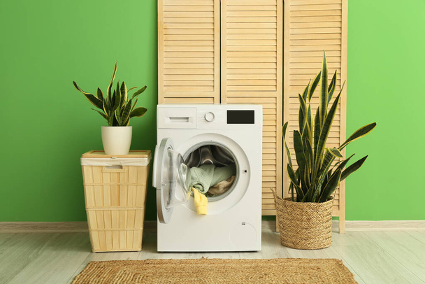 Сучасна пральна машина з білизною, кошиком і рослинами біля кольорової стіни
 - Фото, зображення