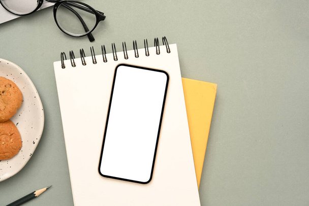 Bovenaanzicht, Minimale werkruimte met smartphone wit scherm mockup op spiraal notitieblok, bril, potlood, en een plaat van cookies op grijze achtergrond. - Foto, afbeelding