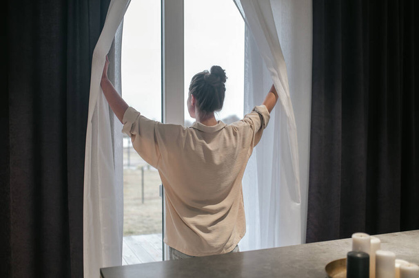 Una mujer con un moño abriendo las cortinas de la habitación - Foto, Imagen