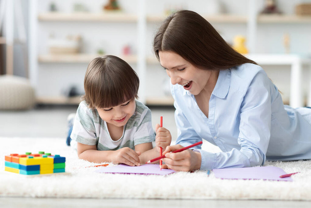 Творческое развитие. Милый счастливый маленький мальчик рисует красочную картину с карандашами, учится с молодой веселой матерью - Фото, изображение