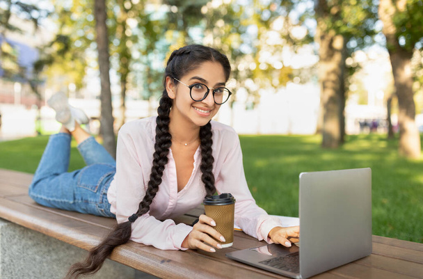 Chica estudiante moderna feliz estudiando al aire libre, descansando con el ordenador portátil y una taza de café para llevar en el banco en el parque - Foto, imagen