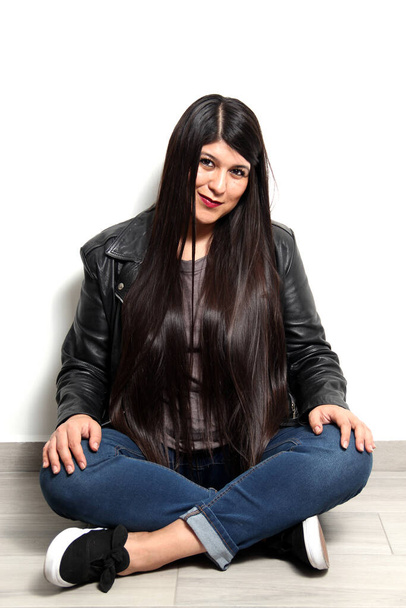 Happy and proud Latin young adult woman shows her long straight, black and shiny hair on a white background with a leather jacket and black tennis shoes - Foto, immagini