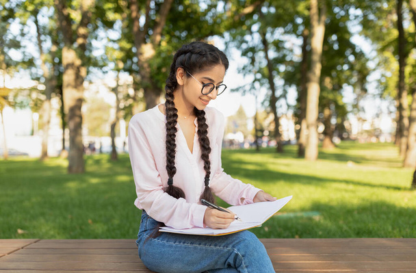Estudiante universitaria escribiendo en cuaderno al aire libre, estudiando y preparándose para el examen mientras está sentada en el banco en el parque - Foto, imagen