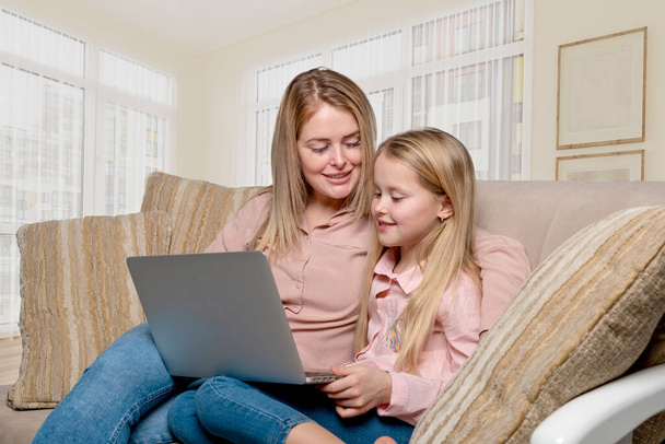 Foto de una mamá y una hija de rubias con el pelo largo con camisas rosas y jeans sentados en el sofá en casa con una computadora portátil.   - Foto, Imagen