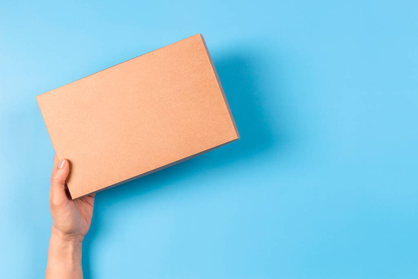 Vista superior a la mano femenina sosteniendo caja de cartón marrón sobre fondo azul claro. Caja de paquetes burlona. Embalaje, compras, concepto de entrega - Foto, Imagen