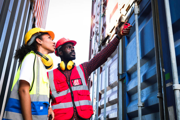 Zwei afroamerikanische Industrieingenieure mit Warnweste und Helm arbeiten zusammen auf dem Containerplatz der Logistikschifffahrt, ein Arbeiter hält Walkie-Talkie in der Hand und zeigt weg. - Foto, Bild