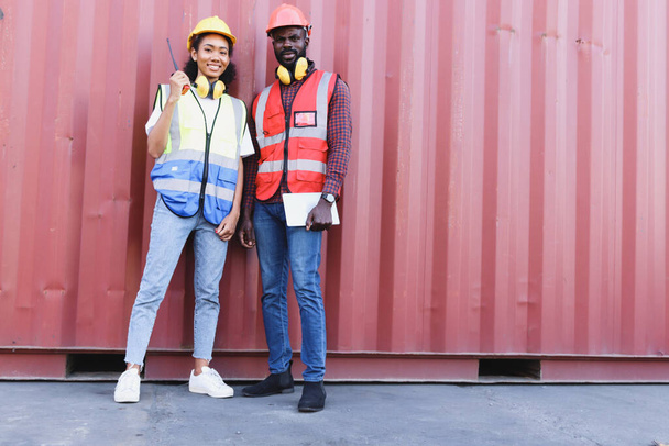 Ritratto di due felici sorridenti ingegnere afroamericano uomo e donna che indossano giubbotto di sicurezza e casco in piedi davanti al container presso il cantiere logistico di trasporto merci. - Foto, immagini