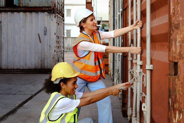 Dvě krásné mladé ženské dělnice nosí záchrannou vestu a helmu, snaží se otevřít lodní kontejnerové dveře v logistickém nákladovém prostoru. Afroameričanky a bělošky spolupracují na stejném pracovišti. - Fotografie, Obrázek