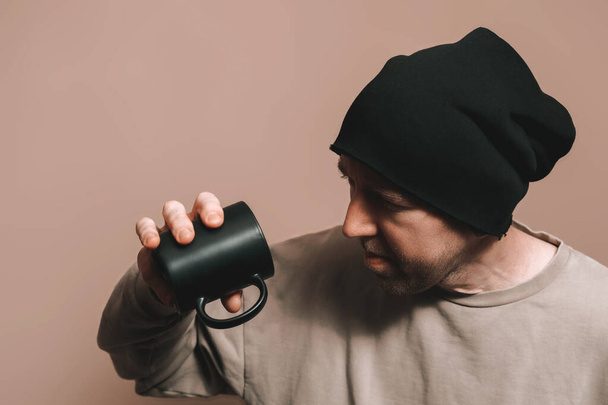 Ein lustiger junger Mann blickt überrascht in eine leere schwarze Tasse. Porträt eines Europäers mit schwarzer Mütze - Foto, Bild