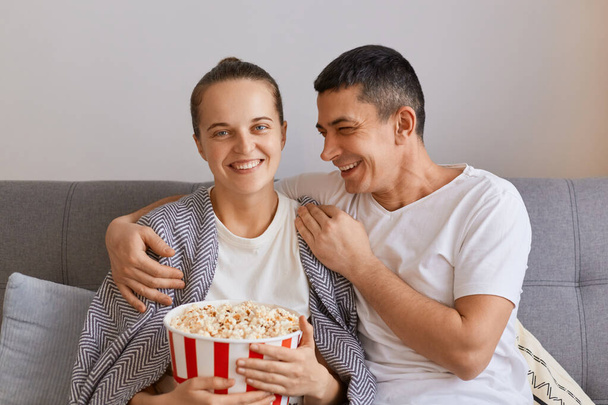 Portrait of smiling joyful wife and husband sitting on sofa with pop-corn and remote control, watching comedy or funny show, laughing happily and hugging, couple spending time together. - Foto, imagen