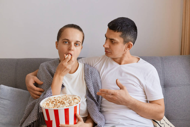 Indoor shot of young couple sitting on sofa with pop-corn and remote control, watching tv or movie and talking, woman with indifferent expression eating snack, man talking to wife. - Fotó, kép