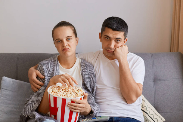 Portrait of bored sad upset wife and husband sitting on sofa with pop-corn and remote control, watching boring movie, looking at camera with frowning faces. - Foto, imagen
