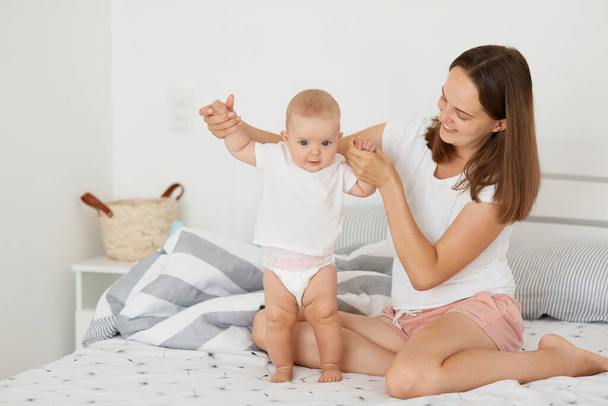 Indoor shot of satisfied young mother wearing white t shirt, attractive woman sitting on bed with her infant daughter, expressing positive emotions, playing at home in bedroom. - Photo, image