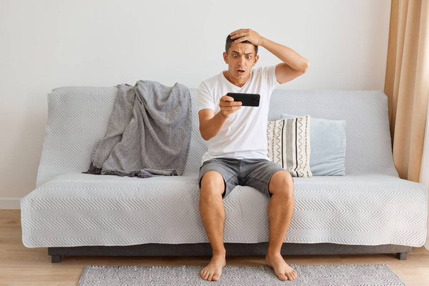 Indoor shot of confused man wearing white t shirt and jeans shirt sitting on sofa at home, holding cell phone in hands, playing online game, loosing the important level. - Foto, afbeelding