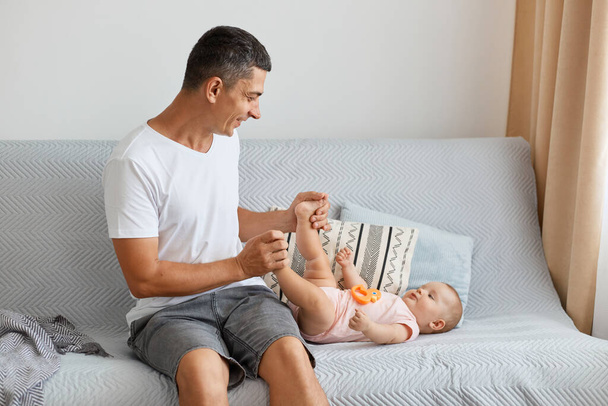 Indoor shot of attractive loving father, handsome man wearing white casual style t shirt sitting on sofa and playing with her infant daughter lying on sofa. - Zdjęcie, obraz