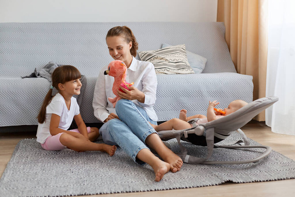 Indoor shot of smiling woman wearing white shirt and jeans sitting on floor near sofa with her daughters, mother showing toy to her elder kid, infant baby lying in rocking chair. - Foto, Bild