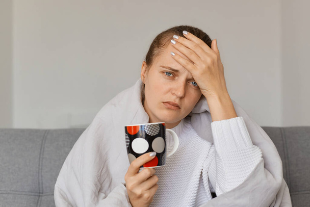 Image of sick ill Caucasian woman being wrapped in blanket sitting on sofa with cup of hot tea, having headache and high temperature, catching cold, feels bad. - Фото, зображення