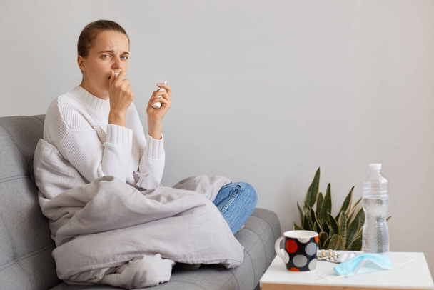 Portrait of sad Caucasian woman being wrapped in blanket sitting on sofa and looking at camera, holding handkerchief in hands, coughing, being infected with covid. - Foto, Bild