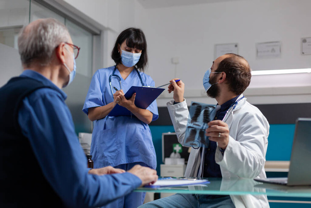Médico hablando con enfermera y sosteniendo resultados de rayos X en visita de chequeo - Foto, imagen