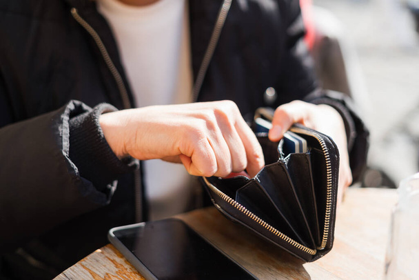 Close up of black purse and woman hands looking for money. Empty purse. Outside sunny day in cafe - Фото, изображение