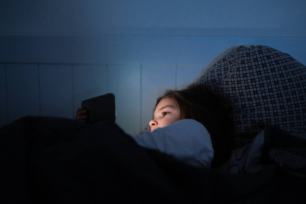 Baby girl is watching video on smartphone in bed before sleeping at evening - Foto, Imagen