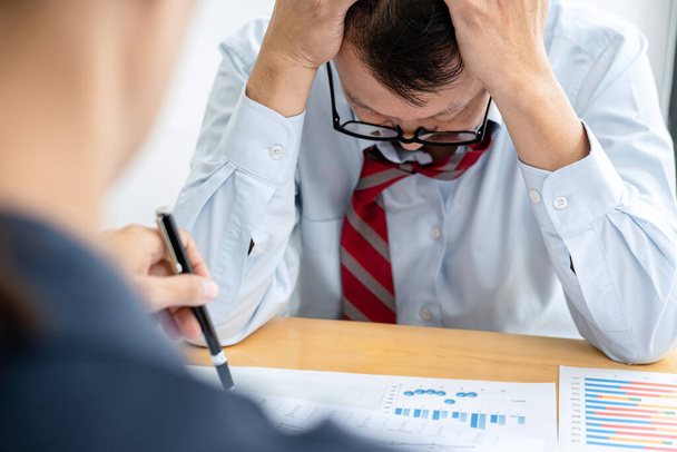 Male employee is holding his head with feeling stress about unsuccessful project after executive checking about business document and complain about the work during meeting in office - 写真・画像