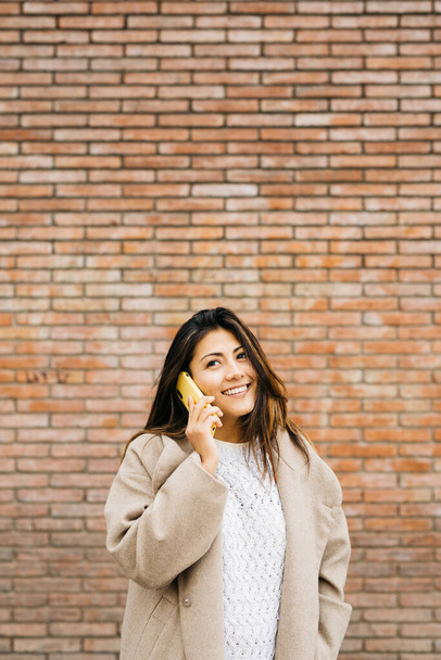 Porträt einer jungen Frau, die ein Telefongespräch führt, vor einer Ziegelwand - Foto, Bild