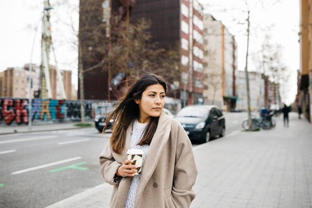 Portret van een jonge vrouw met een koffiekop die op straat loopt - Foto, afbeelding