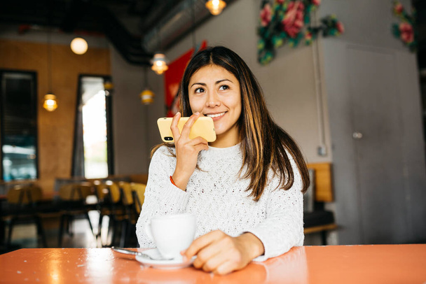Retrato de una joven sonriente usando un teléfono mientras bebe café en una cafetería - Foto, imagen