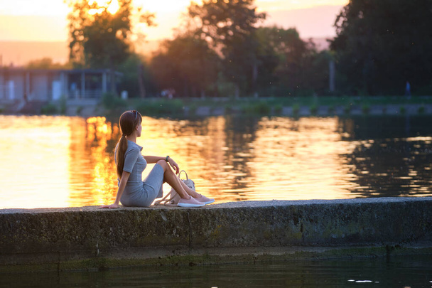 孤独な女性の湖の海岸に暖かい夜に座ってのバックビュー。孤独とリラクゼーションの概念 - 写真・画像