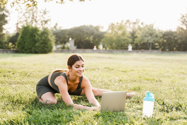 Fit girl with laptop on the green grass. Fitness trainer is chatting online with clients and explain exercises. Sport lifestyle - Photo, Image