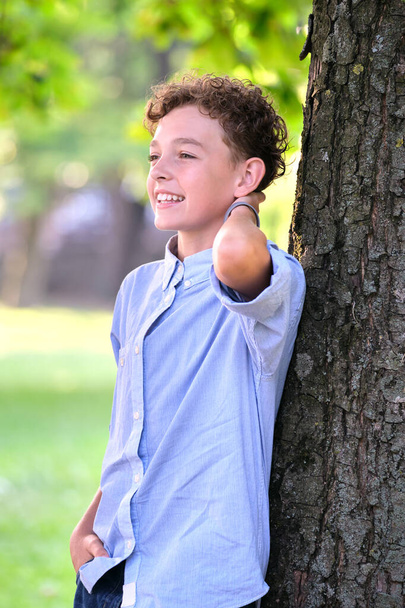 Nuori onnellinen lapsi poika rentoutuu kesäpuistossa nojaten puunrunkoon. Positiivinen nuorukainen nauttii kesästä ulkona. Lastenhyvinvoinnin käsite - Valokuva, kuva