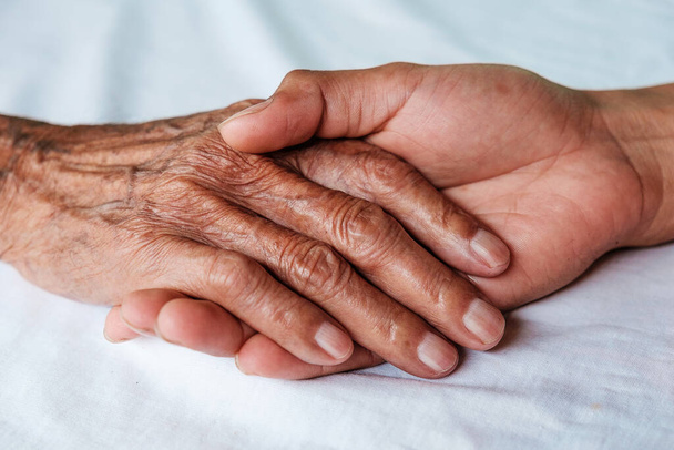Manos del anciano y una mano del hombre sobre la cama blanca en un hospital - Foto, Imagen