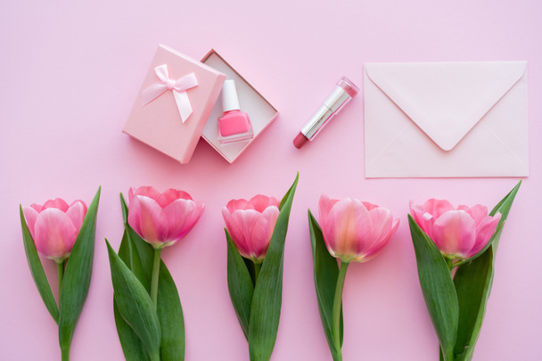 top view of row with blooming tulips near gift box with nail polish near lipstick and envelope on pink - Fotoğraf, Görsel