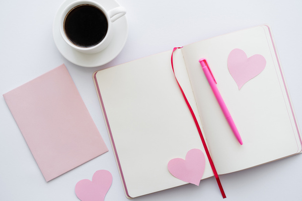 bovenaanzicht van notebook met papieren hartjes en kopje koffie in de buurt van envelop op wit - Foto, afbeelding