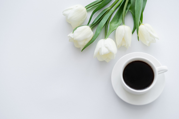 top view of cup with coffee on saucer near tulips on white - Foto, Imagem
