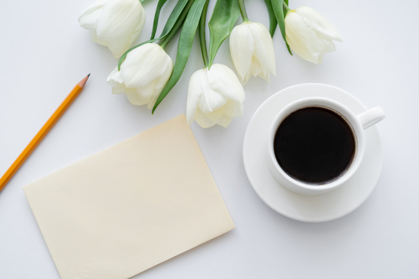horní pohled na obálku s tužkou u šálku s kávou a tulipány na bílém - Fotografie, Obrázek