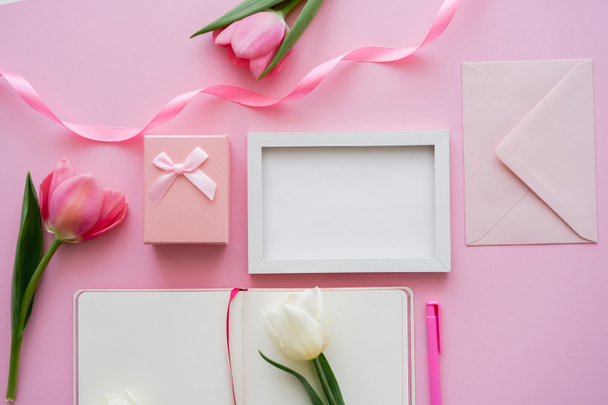 bovenaanzicht van tulpen in de buurt van frame, blanco notitieboekje, envelop en geschenkdoos op roze - Foto, afbeelding