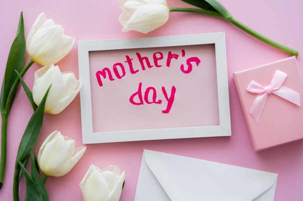 vue du dessus de la carte avec le lettrage de la fête des mères dans le cadre près de la boîte cadeau, enveloppe et fleurs blanches sur rose  - Photo, image