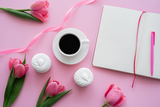bovenaanzicht van kopje met koffie in de buurt van zephyr, blanco notitieboekje en tulpen op roze  - Foto, afbeelding
