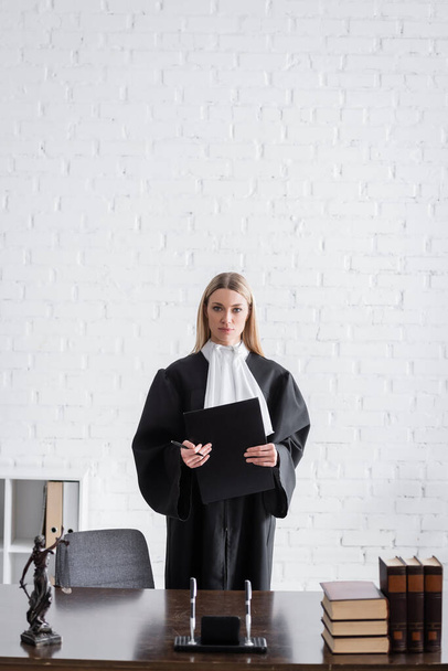 judge in mantle standing with folder near books and themis statue on work desk - Fotoğraf, Görsel