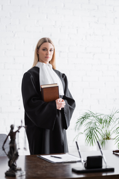prosecutor in black mantle looking at camera while standing with book near white wall - Fotoğraf, Görsel